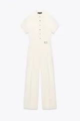 Mono largo color crema, de cuello solapa y manga corta con dobladillo. Tiene bolsillos delanteros y falsos bolsillos traseros. El pantalón es de pierna ancha con pinzas delanteras y dobladillo. Se cierra con cremallera, hebilla y botones.