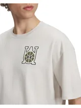 Remera blanca de corte oversize, con cuello redondo y mangas cortas. Presenta un estampado en el lado izquierdo del pecho con el logo de Under Armour y motivos de baloncesto en tonos verde y marrón.