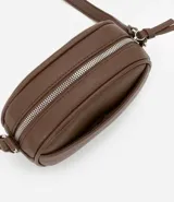 Cartera tipo camera bag pequeña, color marrón camel, de forma ovalada con superficie texturizada. Cuenta con correa ajustable larga, cierre principal con cremallera dorada y un pequeño bolsillo frontal con cremallera.