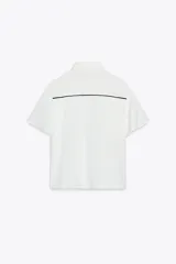 Camisa blanca de corte recto, con cuello solapa y mangas cortas. Presenta vivos negros a contraste en el frente y aberturas laterales en el ruedo. Cierre frontal con botones ocultos.