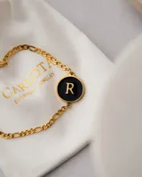 Pulsera dorada con cadena de eslabones y dije redondo negro con la letra R en dorado.