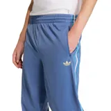 Pantalón deportivo Adidas Firebird de tejido liso, color azul claro, con cintura elástica y tres franjas laterales en tonos de azul y blanco. Presenta un bolsillo lateral con cierre y el logo del trébol dorado bordado en el muslo.