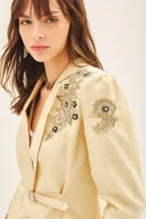 Saco sastre color beige claro, de manga larga y calce al cuerpo, con cierre frontal ajustable mediante cinturón con hebilla de piedras. Presenta bordados florales en tonos dorados y negros con lentejuelas y canutillos en los hombros y solapas.