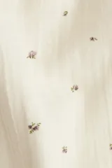 Pijama de dos piezas en muselina de algodón color crema con estampado de pequeñas figuras oscuras. Incluye una camisa de manga larga con cuello camisero, botones frontales y bolsillo de parche, y un pantalón recto con cintura elástica y cordón de ajuste.