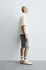 Camisa de denim de algodón, color beige claro, con corte relajado, cuello bowling y manga corta. Presenta cierre frontal con botones a presión y un pequeño bordado a tono en el pecho.