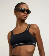 Top deportivo negro sin costuras, con escote asimétrico y doble tirante en los hombros. Presenta una delgada línea blanca horizontal en la banda inferior.