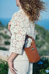 Cartera tipo balde color marrón con asa de mano rígida y correa bandolera ajustable y extraíble. Cierre con trabilla y adorno metálico dorado.