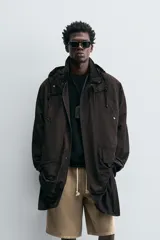 Trench relaxed fit negro confeccionado en tejido técnico ligeramente acolchado, con cuello subido y capucha desmontable. Tiene manga larga, bolsillos de vivo en pecho y de solapa en cadera, y abertura en el centro de la espalda. El cierre frontal es con cremallera oculta por solapa con botones a presión.
