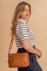 Bandolera de simil cuero color marrón topo, con textura ligeramente desgastada y detalles en nobuck. Presenta múltiples bolsillos frontales con cierres metálicos y tiradores de cuero. Correa ajustable para llevar al hombro o cruzada.