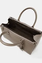 Bolso de mano tipo satchel o tote pequeño, color topo o gris pardo, con doble asa superior y cierre principal por cremallera. Presenta un colgante rectangular de cuero sintético colgando de una de las asas.