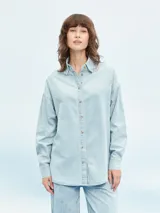 Camisa celeste de denim con botones marrones, corte holgado y mangas largas.