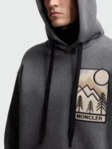 Buzo con capucha gris de Moncler, con cordones negros y parche bordado en el pecho con diseño de paisaje montañoso y logo de la marca.