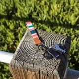 Llavero de muñeca con cinta textil estampada a rayas horizontales en tonos pastel y oscuros (rosado, beige, verde azulado, naranja). Incluye un detalle de cuero sintético marrón con logo grabado y un gancho metálico tipo mosquetón de color bronce envejecido.