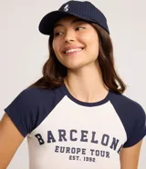 Remera ajustada al cuerpo, estilo raglán, con cuerpo blanco y mangas cortas azul marino. Presenta un estampado en el pecho con el texto "BARCELONA EUROPE TOUR EST. 1992" en azul marino.