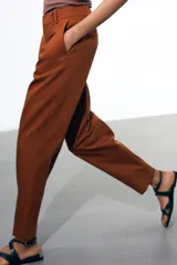 Pantalón color marrón confeccionado en hilatura con mezcla de lyocell y algodón. Tiro medio, cintura con trabillas, bolsillos laterales, pinzas en delantero y raya planchada. Cierre frontal con cremallera, botón y ganchos metálicos.