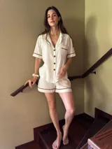 Pijama de algodón color blanco, compuesto por una camisa de manga corta abotonada con cuello solapa y un bolsillo en el pecho, y un short con elástico en la cintura. Ambas prendas tienen un detalle de picot marrón en los bordes.