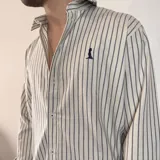 Camisa de manga larga color beige claro, de corte regular, con cuello clásico y cierre frontal con botones. Presenta un pequeño logo bordado en el pecho.