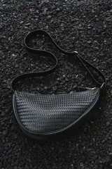 Cartera bandolera negra con diseño trenzado, forma asimétrica, cierre superior y correa ajustable.