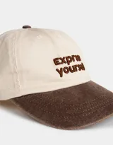 Gorra de béisbol color beige con visera marrón y bordado frontal con la frase "Express Yourself" en marrón. Cuenta con botón superior marrón y regulador trasero metálico.