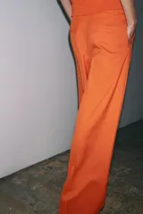 Pantalón ancho color naranja, de tiro medio y cintura con trabillas. Tiene bolsillos laterales y de vivo en espalda, pinzas en delantero, raya planchada y cierre frontal con cremallera, botón y ganchos metálicos.