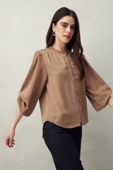 Camisa color marrón claro con cuello mao, mangas 3/4 abullonadas y cierre frontal con botones. Presenta pequeños pliegues en los hombros y detalle calado sutil.