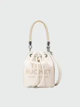 Bolso tipo bucket de cuero color gris con logo "The Bucket Marc Jacobs" estampado en negro. Tiene asa corta de cuerda trenzada y correa larga de cuero ajustable y removible.