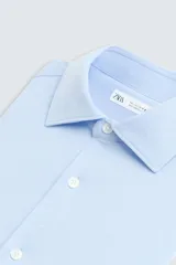 Camisa celeste a rayas verticales blancas, de corte recto, con cuello italiano abierto, mangas largas con puños abotonados y cierre frontal con botones.