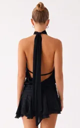 Vestido corto negro con escote halter ajustable con lazo en el cuello y espalda abierta con tira regulable. Presenta cintura fruncida con efecto fajín y falda corta con volados. El tejido es satinado con textura de rayas sutiles.