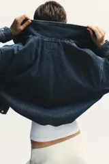 Chaqueta corta de denim azul con cuello alto, manga larga con hombros caídos, bolsillos laterales, bajo con elástico y cierre frontal con cremallera y botones ocultos.