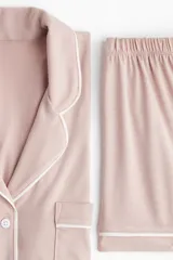 Pijama corto de dos piezas color rosado, en tejido de punto suave con ribetes blancos en contraste. Incluye camisa de manga corta con cuello abierto, botones al frente y bolsillo superior, y short con cintura elástica.