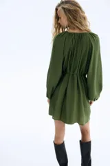 Vestido corto color verde oliva, de corte holgado, con cuello redondo fruncido, mangas globo largas con puños elásticos y botonadura frontal. Incluye cinturón fino para atar.
