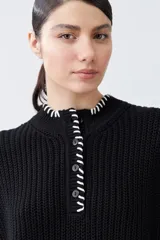 Chaleco negro de tejido de punto con cuello alto, cierre parcial con botones y detalle de pespunte blanco en el ruedo y el cuello. Sin mangas.
