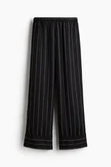 Pantalón holgado negro con finas rayas verticales blancas, tiro alto, cintura elástica, bolsillos laterales y ribete decorativo en los bajos.