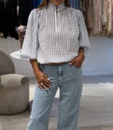 Blusa a rayas verticales celestes y blancas, de manga larga con puños elásticos, cuello alto con volantes y cierre frontal parcial con botones.