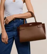 Cartera satchel mediana color marrón, confeccionada en cuero sintético con detalles en gamuza. Cuenta con asas medianas, cierre de cremallera y detalles metálicos dorados. Incluye correa extraíble y regulable.