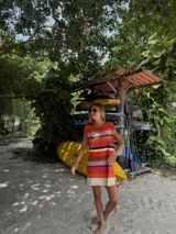 Vestido corto tejido con hilo de algodón a rayas horizontales en tonos celeste, azul, marrón, blanco, naranja y verde. Tiene cuello redondo y mangas cortas. El ruedo es desflecado.