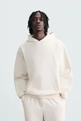 Sudadera con capucha color crudo, de corte relajado, confeccionada en tejido de algodón con interior rizado. Cuenta con cuello con capucha, manga larga, bolsillo frontal tipo canguro y acabados en rib.