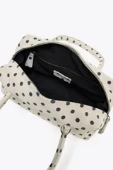Bolso tipo bowling color crudo con estampado de lunares negros. Tiene doble asa de mano y correa bandolera extraíble. Incluye bolsillo interior con cremallera y cierre principal con doble cremallera.