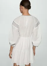 Vestido corto blanco de algodón con bordados y detalles calados. Tiene cuello redondo, mangas largas con puños elásticos, cintura elástica con lazo y diseño evasé.