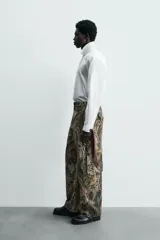 Camisa blanca de algodón popelín, corte relajado, con cuello solapa y pechera frontal plisada. Las mangas terminan en puños ajustables con lazos largos estampados en color marrón con patrón circular.