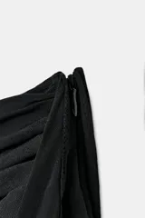 Falda pantalón negra de tiro bajo con cintura drapeada y bajo acabado en volantes superpuestos. Presenta un lazo largo y fino que cae desde el lateral.