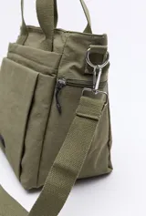 Cartera de nylon color verde militar con asas superiores reforzadas y correa larga regulable y desmontable. Cuenta con bolsillos frontales y laterales con cierre metálico, y un logo de la marca en la parte inferior central.