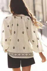 Blusa color crudo de cuello redondo con puntilla, frunces y cordón. Escote pico y manga larga abullonada. Detalle de bordados a contraste en negro.