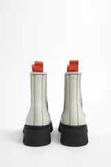 Botines Chelsea de cuero sintético blanco con puntera y suela track de goma negra. Elásticos laterales negros y tirador trasero naranja.