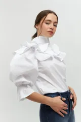 Camisa blanca de algodón con mangas abullonadas y puños fruncidos. Presenta un cuello removible tipo babero con volantes estructurados y tablones, y un lazo largo que puede anudarse al frente.