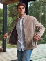 Campera bomber color beige, de corte clásico, con cuello redondo, cierre frontal con cremallera, bolsillos laterales con cremallera y bolsillo en la manga.