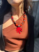 Collar corto con cuentas de coral, cerámica y acrílico en tonos naranja, rojo, marrón y verde. Dije de hoja pintado a mano en color rojo. Cierre con cadena de acero quirúrgico y ganchito para regular el largo.