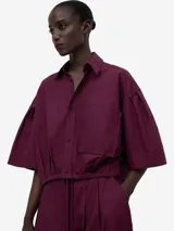 Camisa boxy color borgoña de algodón orgánico, con cuello solapa, manga corta sobre el codo, botonadura frontal, bolsillo en el pecho, canesú en la espalda y ceñidor con cordón en el bajo.
