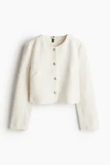 Chaqueta corta color crema en tejido bouclé, de corte entallado, con escote redondo, cierre frontal con botones dorados y forro interior.