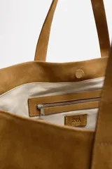 Bolso tipo shopper color marrón claro, fabricado en piel con acabado serraje. Tiene un compartimento principal con cierre imantado y un bolsillo interior con cremallera. Cuenta con dos asas de mano fabricadas en piel.
